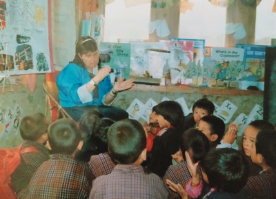 A woman sits in a chair and a classroom of children sit on the floor around her